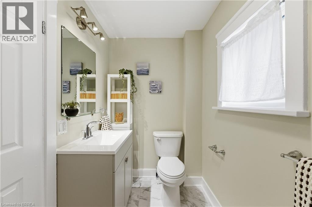 Vanity with integrated sink and chrome faucet, wall-mounted mirror, track lighting, white porcelain toilet, and window with white trim - 87 Strathcona Avenue, Brantford, ON