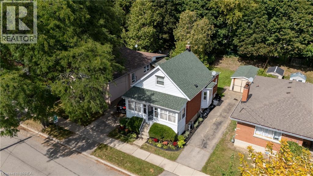 White siding exterior with brick accents, green shingle roof, and a mature tree canopy - 87 Strathcona Avenue, Brantford, ON