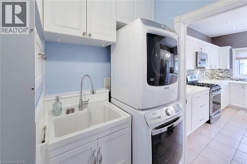 Utility sink with chrome faucet, upper and lower cabinetry, stackable washer and dryer, and tiled flooring - 214 Cloverleaf Drive, Ancaster, ON