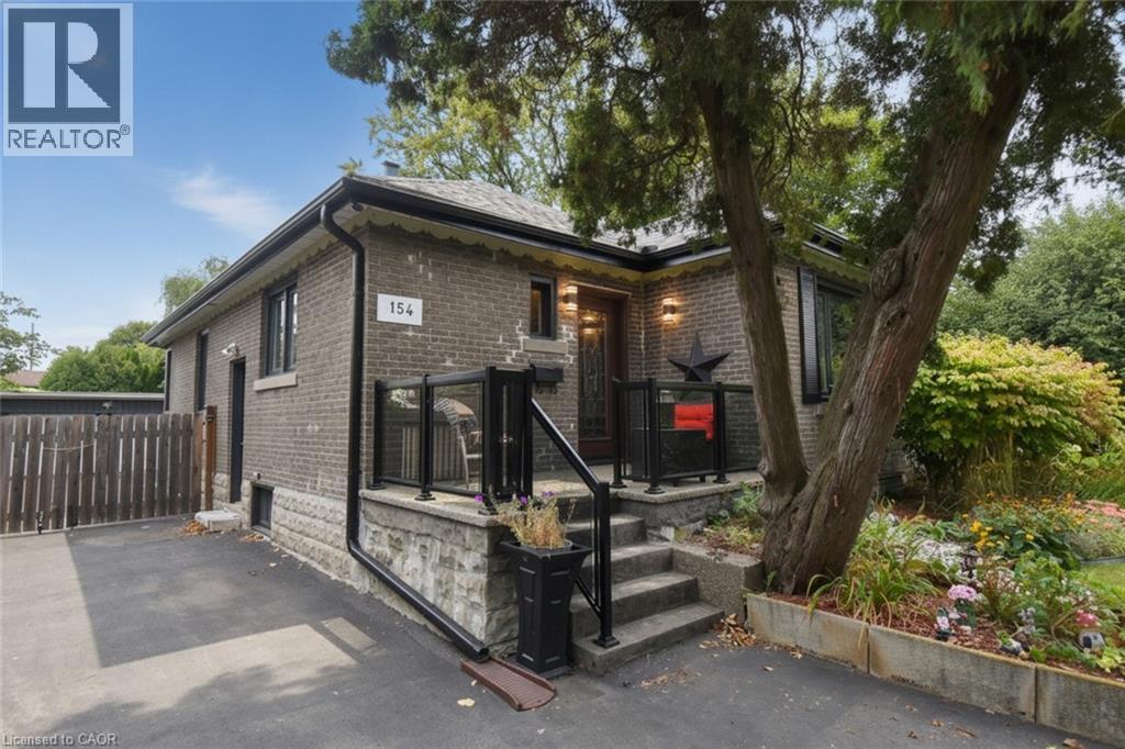 Brick exterior with stone foundation detailing, featuring a paved driveway, front porch with glass railings, and a mature tree - 154 Ewen Road, Hamilton, ON