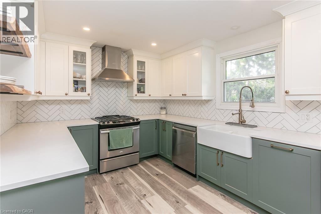 Modern kitchen featuring two-tone cabinetry, stainless steel appliances, a white farmhouse sink, herringbone tile backsplash, and light wood-finish flooring - 393 Huron Street, New Hamburg, ON