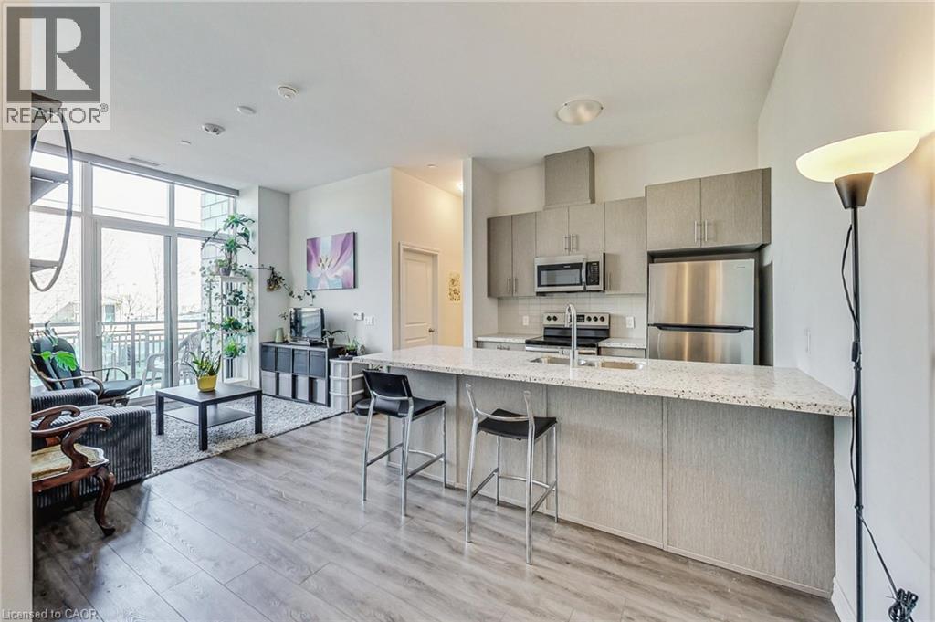 Open-concept living space featuring wood-finish flooring, a kitchen island with seating, stainless steel appliances, and a full-height window with a balcony - 2750 King Street E Unit# 103, Hamilton, ON