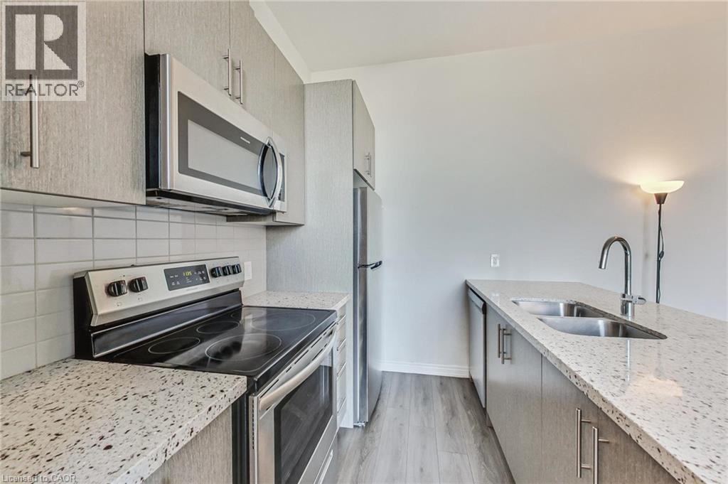 Modern kitchen featuring stainless steel appliances, speckled countertops, a tile backsplash, wood-finish flooring, and light-toned cabinetry - 2750 King Street E Unit# 103, Hamilton, ON