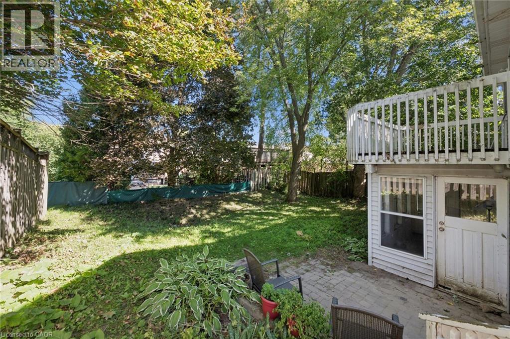 Expansive green space featuring mature trees, a wooden privacy fence, a brick paver patio, an exterior door, and an upper-level balcony with white railings - 512 Thorndale Drive, Waterloo, ON