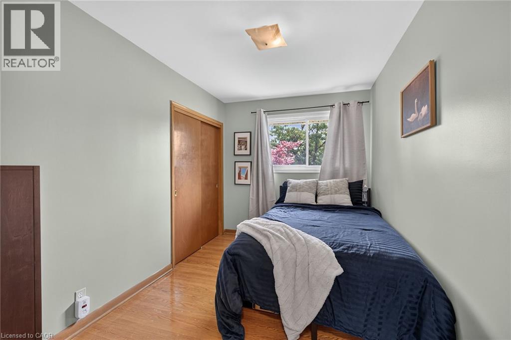 Bedroom featuring wood-finish flooring, a double-door closet with wood panels, and a window with a treed outdoor scene - 13 Allanburg Road S, St. Catharines, ON
