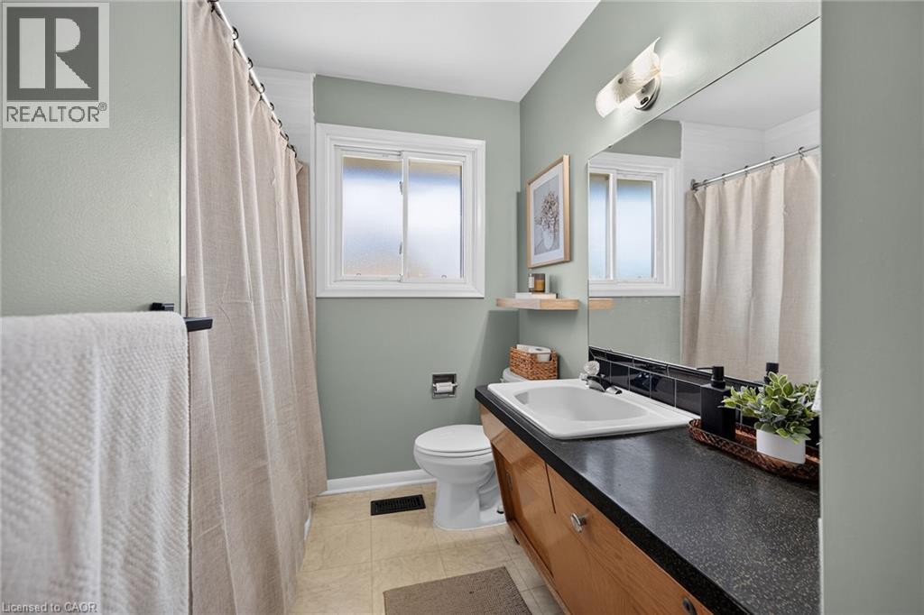 Spa-inspired bathroom featuring soft sage-green walls, dual vanity with wood-finish cabinetry and dark countertop, rectangular vessel sink, chrome fixtures, and bright natural light from dual windows - 13 Allanburg Road S, St. Catharines, ON