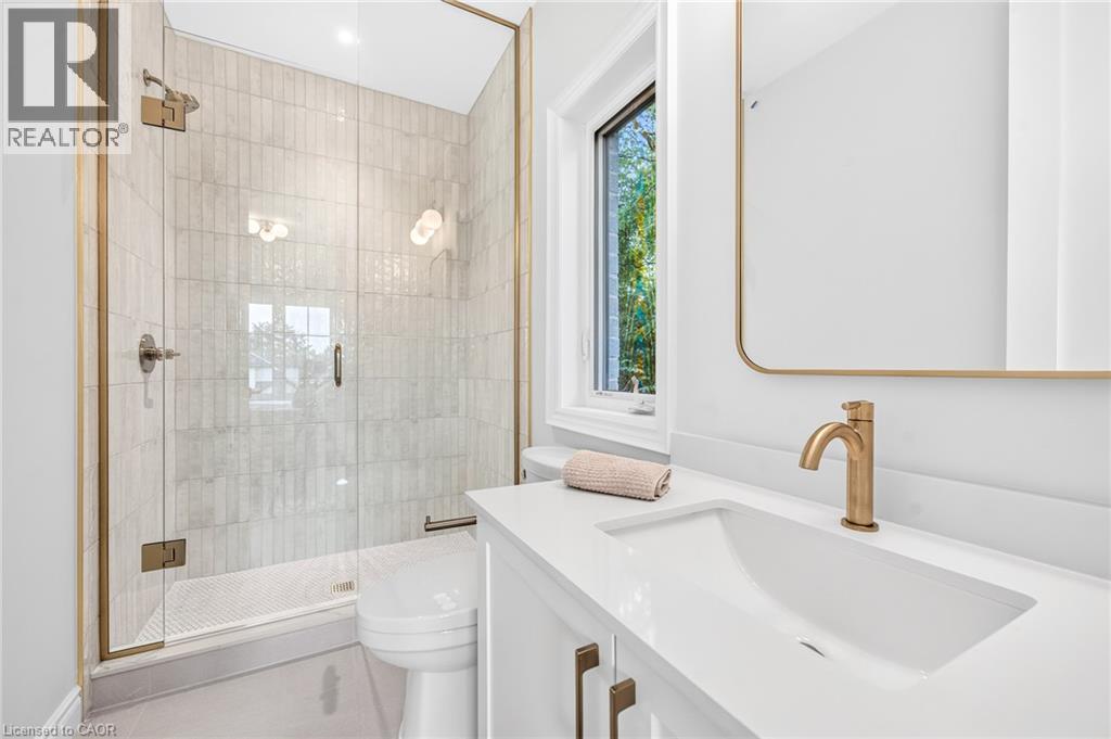 Walk-in shower with clear glass enclosure and gold-toned fixtures, rectangular wall-mounted mirror, single vanity with integrated sink, gold-toned faucet, and white countertop - 67 Cielo Court, Hamilton, ON
