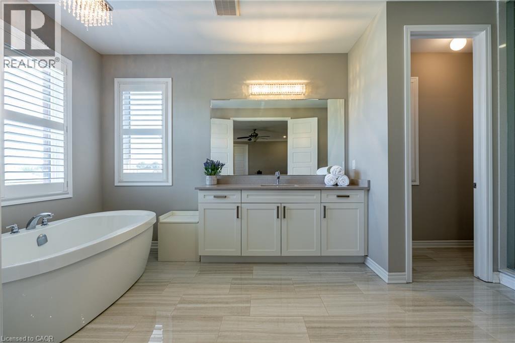 Freestanding soaking tub, white plantation shutters, dual-sink vanity with light-toned countertop, large wall-mounted mirror, and tile flooring - 455 Equestrian Way, Cambridge, ON