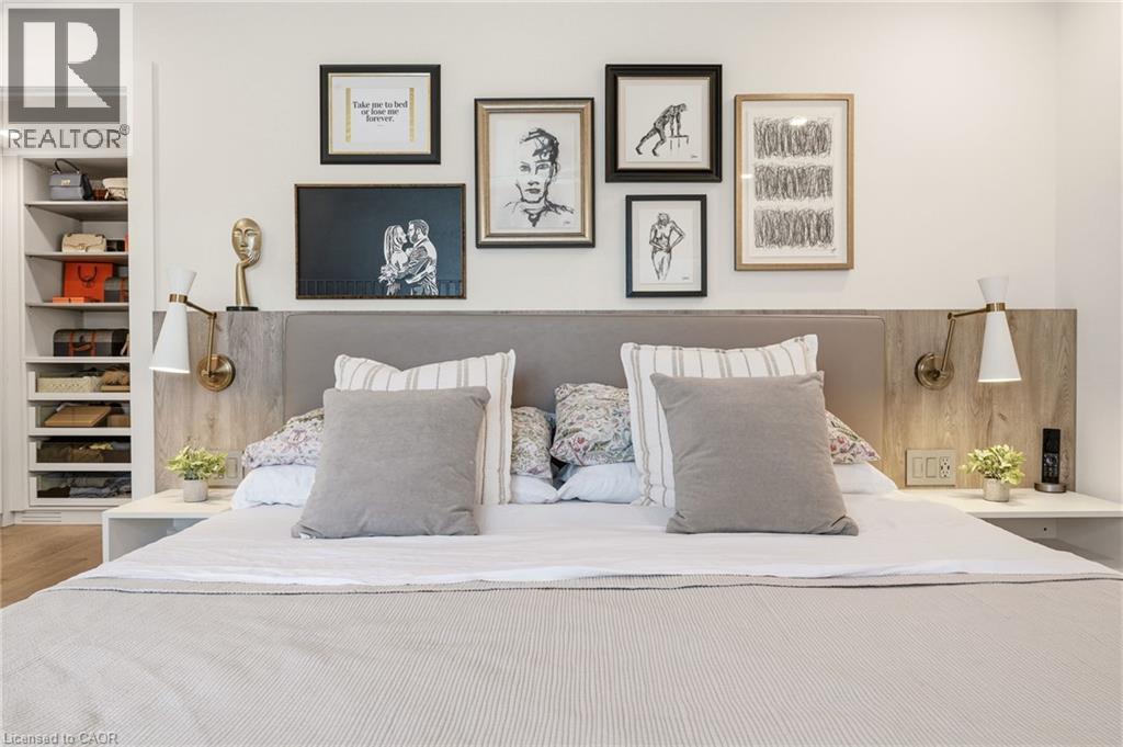 Built-in headboard featuring a fabric-padded panel and wood-finish side panels, integrated wall sconces, floating bedside tables, and a walk-in closet with built-in shelving - 5056 New Street Unit# 4, Burlington, ON