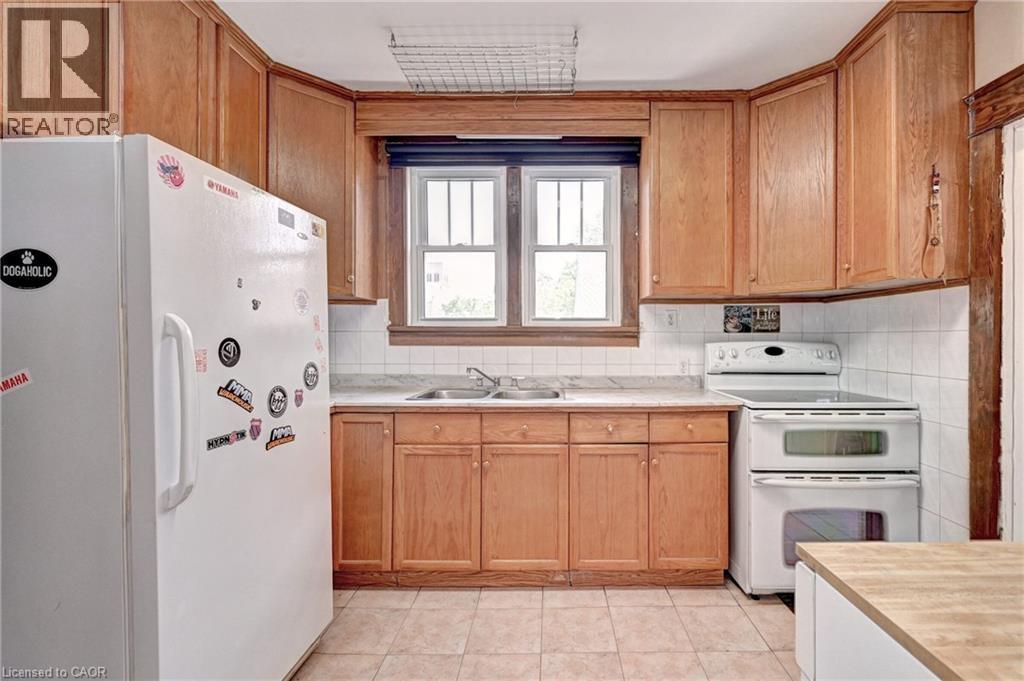 Kitchen featuring a double basin sink, white tile backsplash, wood-finish cabinetry, and neutral-toned floor tiling - 147 Courtland Avenue E, Kitchener, ON - Indoor Photo Showing Kitchen With Double Sink