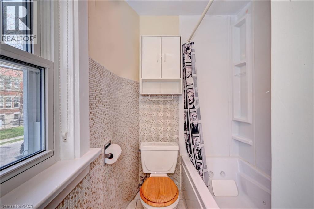 Bathroom featuring a white toilet with a wood-finish seat, mosaic tile wall, a built-in wall cabinet, and a white tub with integrated shelving - 147 Courtland Avenue E, Kitchener, ON - Indoor Photo Showing Bathroom