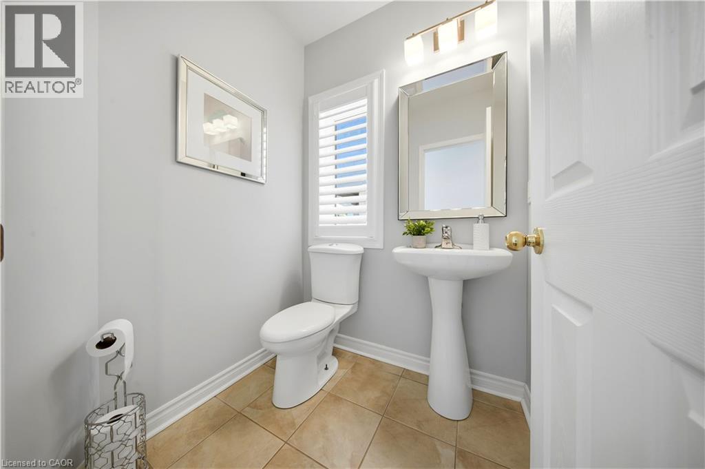 Pedestal sink with chrome faucet, wall-mounted mirror, window with white louvered blinds, tile flooring, and a two-piece toilet - 3352 Hiscott Avenue, Burlington, ON