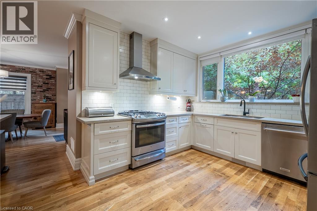 Kitchen featuring wood-finish flooring, white cabinetry, stainless steel appliances, subway tile backsplash, and a large window - 283 Plains Road W, Burlington, ON