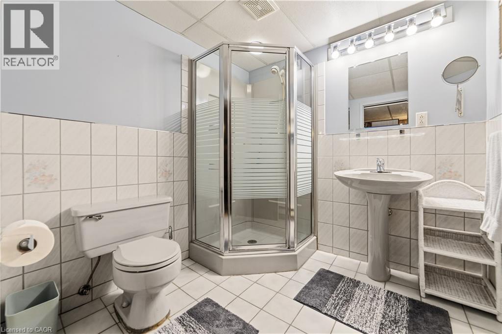 Bathroom in basement with a corner glass-enclosed shower, grey pedestal sink with matching toilet, and light grey ceramic tile flooring - 450 Brigadoon Drive, Hamilton, ON