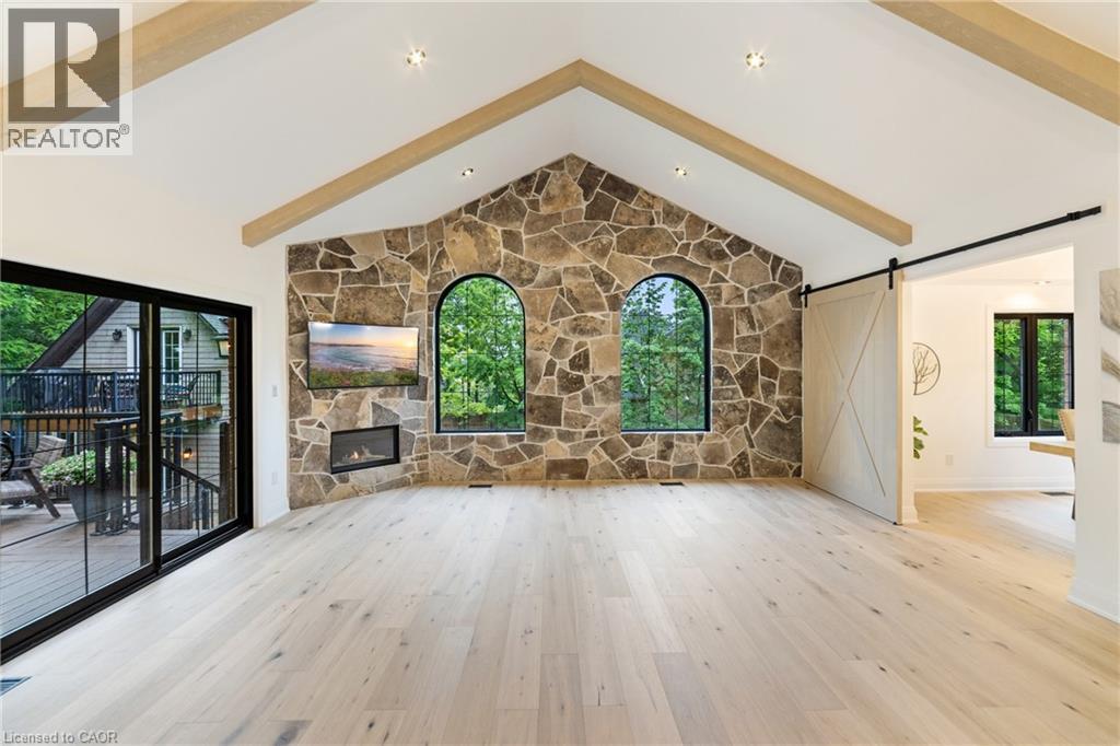 Living room featuring luxury flooring, a barn door to the dining room, gas fireplace, beam ceilings and stone wall, sliding glass doors leading onto expansive 40'x16' composite deck - 515 Hamilton Drive, Ancaster, ON
