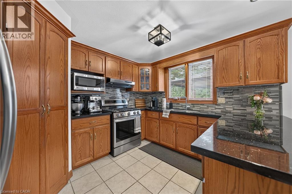 Kitchen with wood cabinetry, black countertops, stainless steel appliances, a mosaic tile backsplash, and a ceiling-mounted light fixture - 52 Archwood Crescent, Cambridge, ON