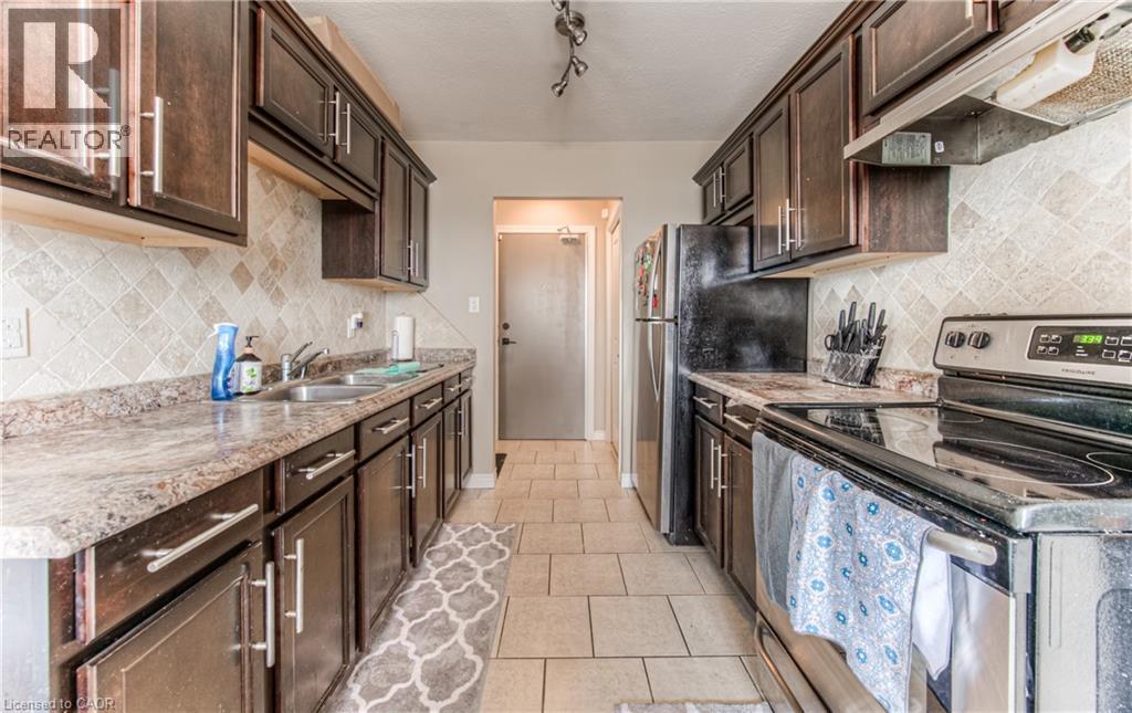 Kitchen featuring stainless steel electric range oven, under cabinet range hood, dark brown cabinets, decorative backsplash, and light tile patterned floors - 283 Fairway Road N Unit# 501, Kitchener, ON