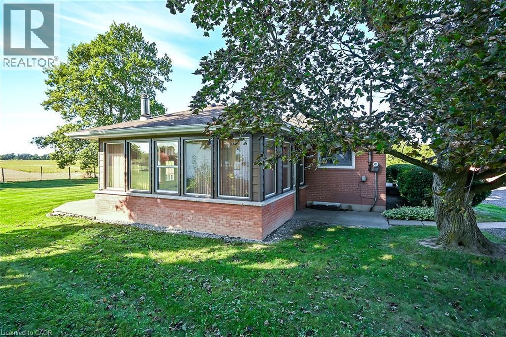 Brick exterior with a sunroom featuring multiple windows, a concrete patio, and a grass lawn with mature trees - 799 Highway 6, Caledonia, ON