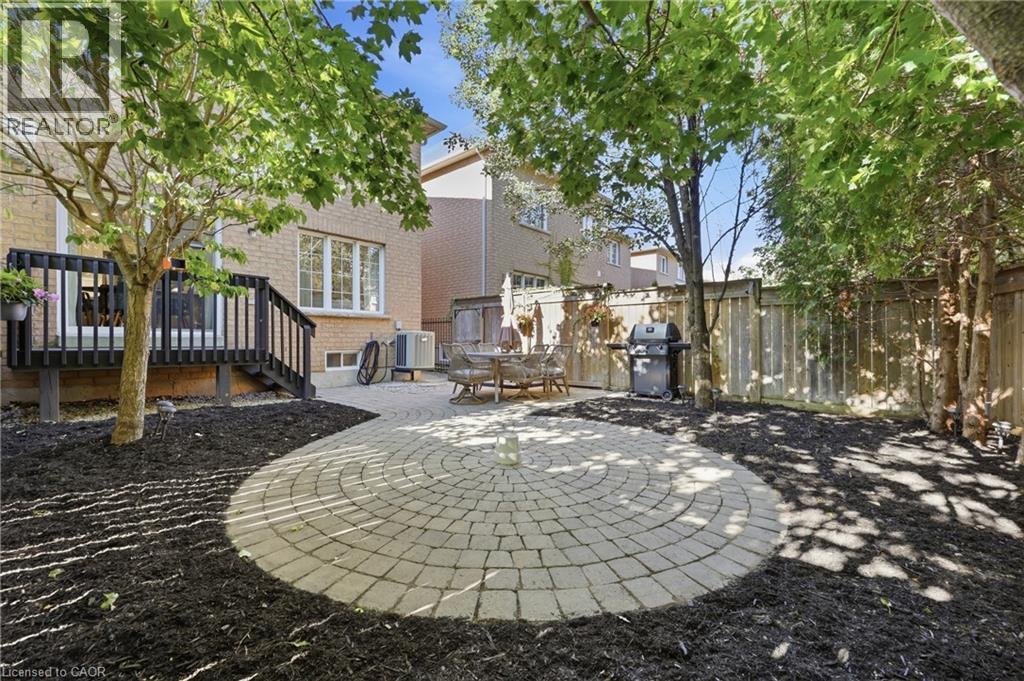 Circular paver patio, wood deck with black railing, mature trees, wood fence, and brick exterior - 2409 Postmaster Drive, Oakville, ON