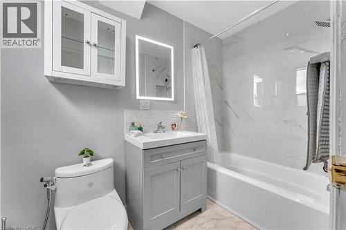 White wall-mounted cabinet with glass-front doors, illuminated mirror, gray vanity with integrated sink, white tile backsplash, and a tub with white subway tile surround - 101 Emerald Street N, Hamilton, ON 