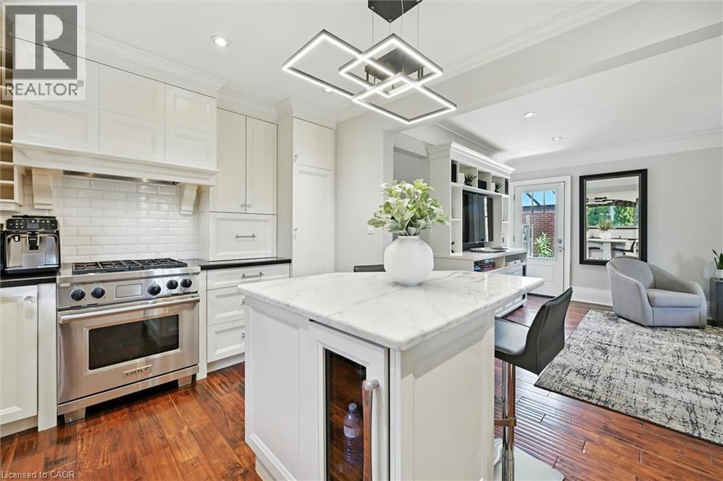 Kitchen featuring white shaker-style cabinetry, a stainless steel range with a tile backsplash, wood-finish flooring, a kitchen island with a beverage cooler, and a contemporary ceiling light fixture - 2209 Ghent Avenue, Burlington, ON