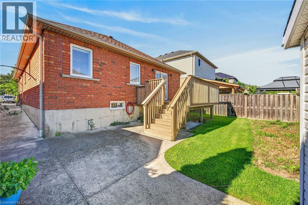 Brick exterior with white frame windows, concrete patio, elevated wood deck with stairs, and a fenced yard with a grass lawn - 795 Tenth Avenue, Hamilton, ON
