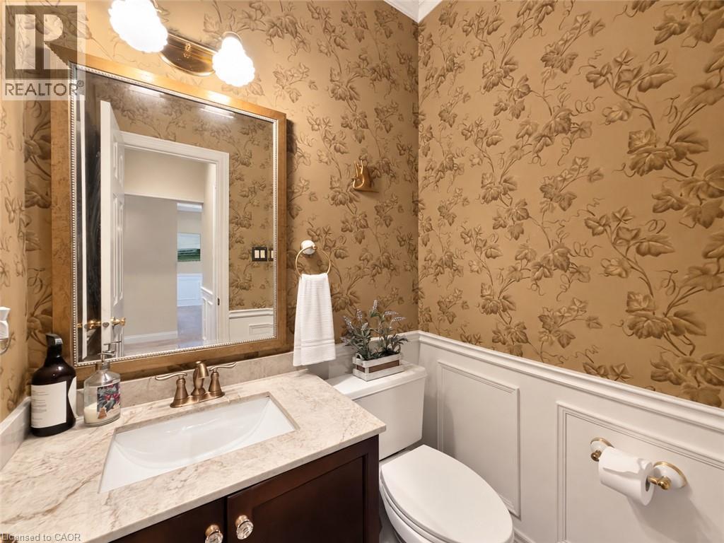 Powder room featuring botanical wallpaper, wainscoting, an undermount sink, a stone-finish countertop, and a wood-finish vanity - 74 Davidson Boulevard, Dundas, ON