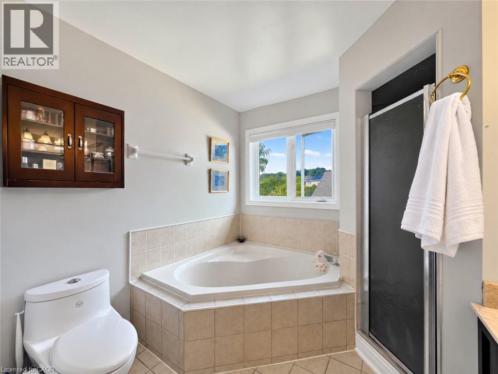 Corner soaking tub with tile surround, walk-in shower with sliding door, wall-mounted wood-finish cabinet with glass doors, and a white toilet - 74 Davidson Boulevard, Dundas, ON