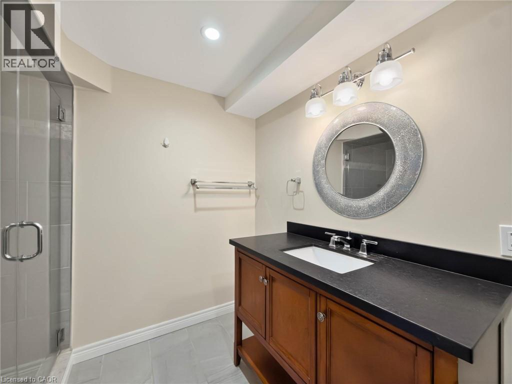 Glass-enclosed shower with tile surround, wood-finish vanity with an undermount sink, dark countertop, and decorative circular mirror - 74 Davidson Boulevard, Dundas, ON