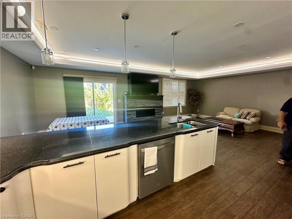 Kitchen island with dark countertop and integrated sink, stainless steel dishwasher, white cabinetry with dark pulls, wood-finish flooring, and recessed lighting - 5 Arden Avenue, Hamilton, ON