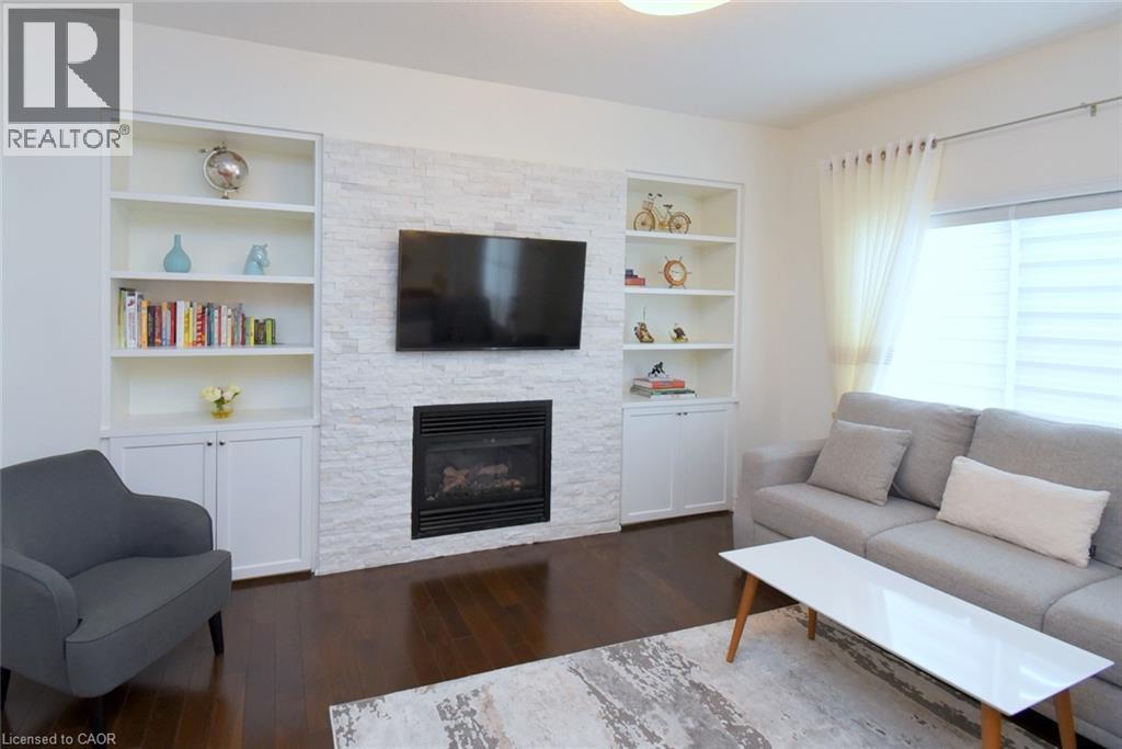 Bright, open living room featuring a white brick fireplace with built-in shelving and cabinetry, wall-mounted flat-screen television, wood-finish flooring, and abundant natural light from multiple win - 25 Guest Street, Hamilton, ON
