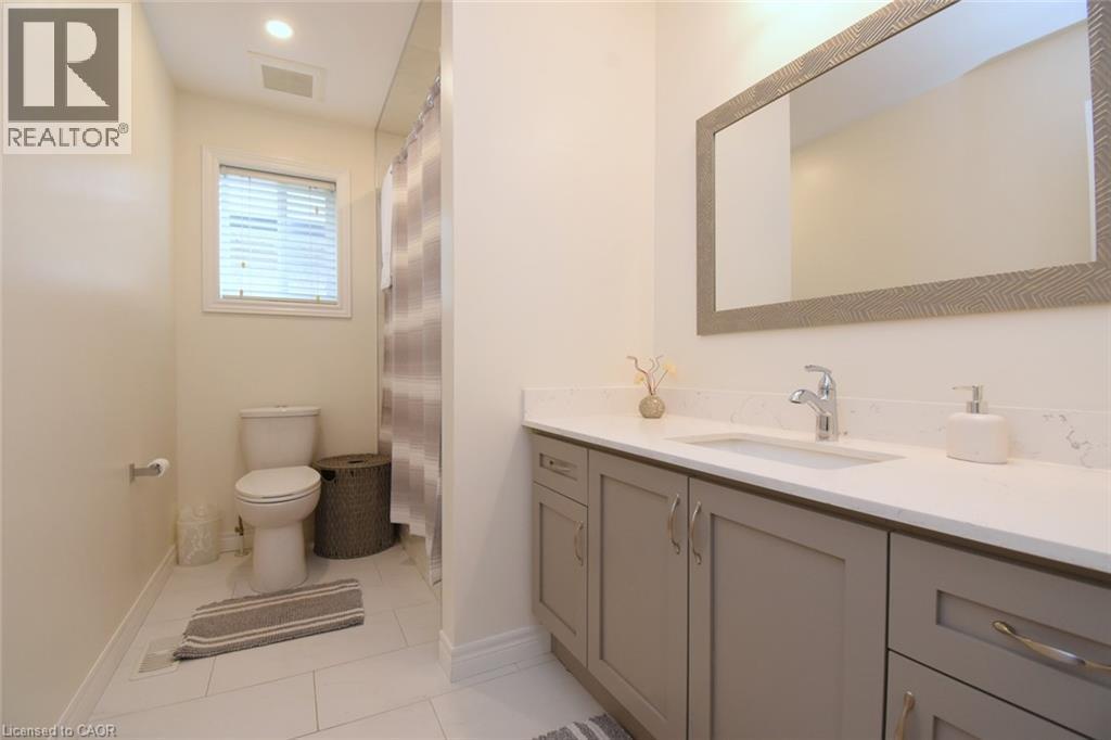 Rectangular mirror with textured frame, single basin vanity with white countertop, chrome faucet, shaker-style cabinetry, and white tile flooring - 25 Guest Street, Hamilton, ON