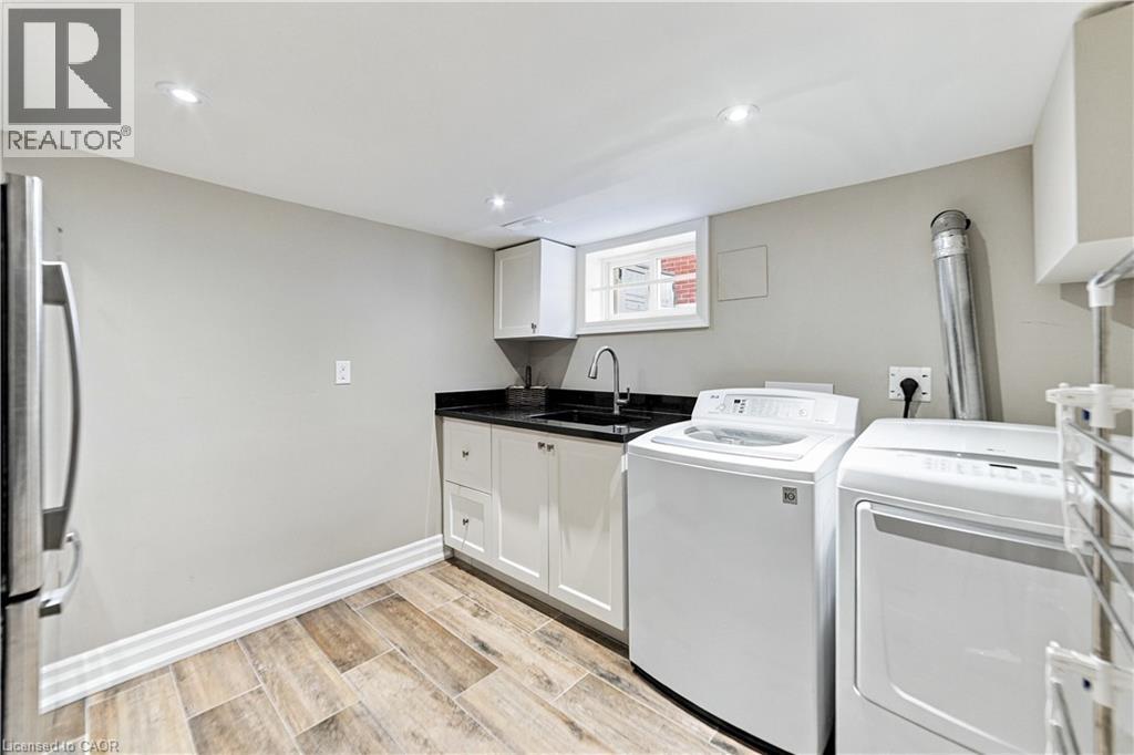 White shaker cabinetry with a dark countertop, stainless steel sink, and wood-finish ceramic flooring. Washer, dryer and fridge included. - 58 Uplands Avenue, Hamilton, ON