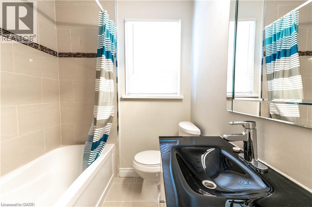 Porcelain bathtub with tiled surround featuring an accent border, black countertop with an integrated sink, and a wall-mounted mirror - 499 East 16Th Street, Hamilton, ON