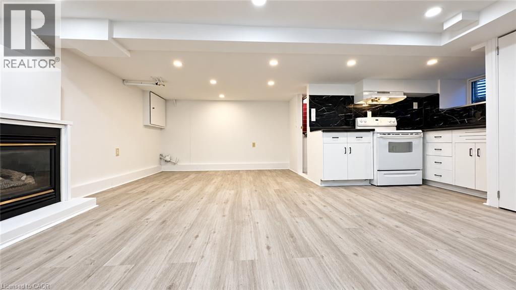 Recessed lighting illuminates the open-concept space featuring wood-finish flooring, a contemporary fireplace, and a kitchen area with white cabinetry, a white range, and a dark stone-look backsplash - 76 Avenue Road, Cambridge, ON