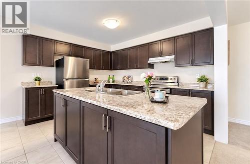 Kitchen featuring dark wood-finish cabinetry, light-toned countertops, stainless steel appliances, a center island with an integrated sink, and tile flooring - 61 Duckworth Road, Cambridge, ON 
