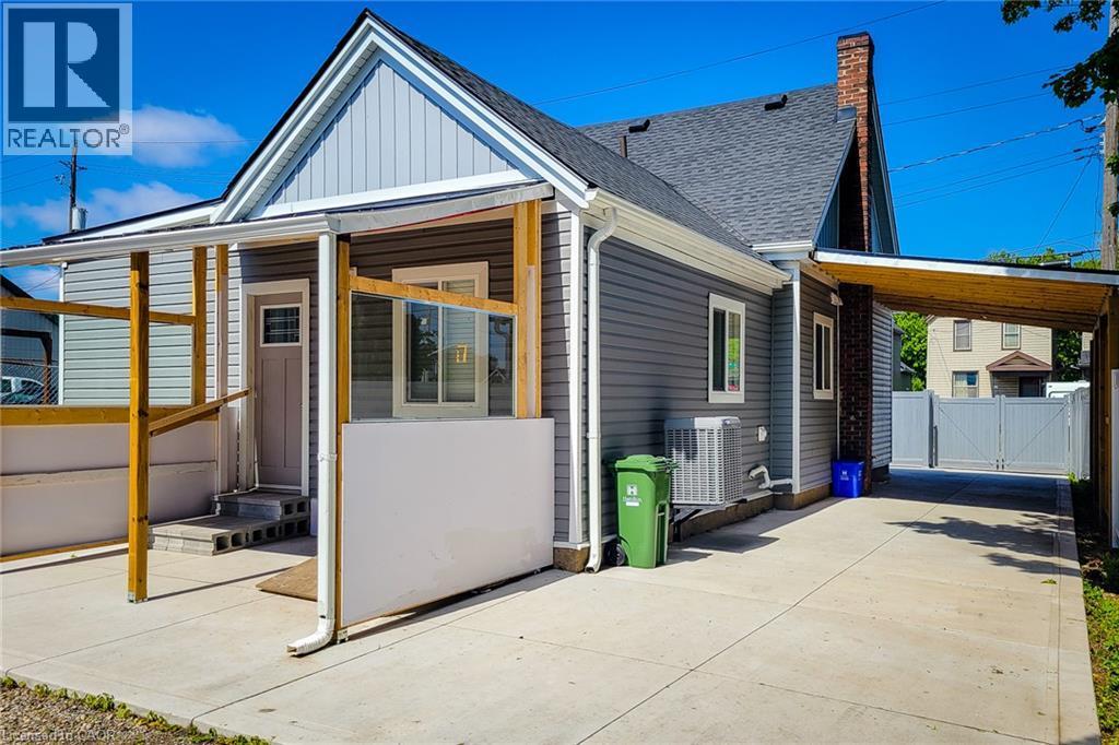 Grey horizontal siding, dark shingle roof, covered patio with wooden supports, concrete driveway, and brick chimney - 78 Hillyard Street, Hamilton, ON