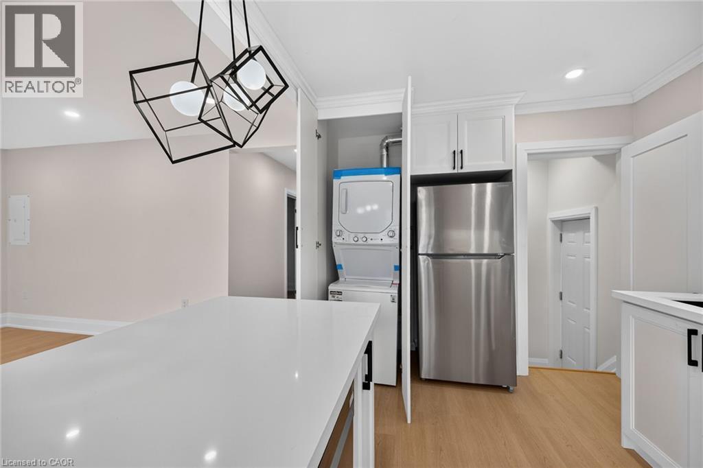 White quartz countertop, wood-finish flooring, white cabinetry with black hardware, stainless steel refrigerator, and stacked washer/dryer units - 50 West 4Th Street, Hamilton, ON
