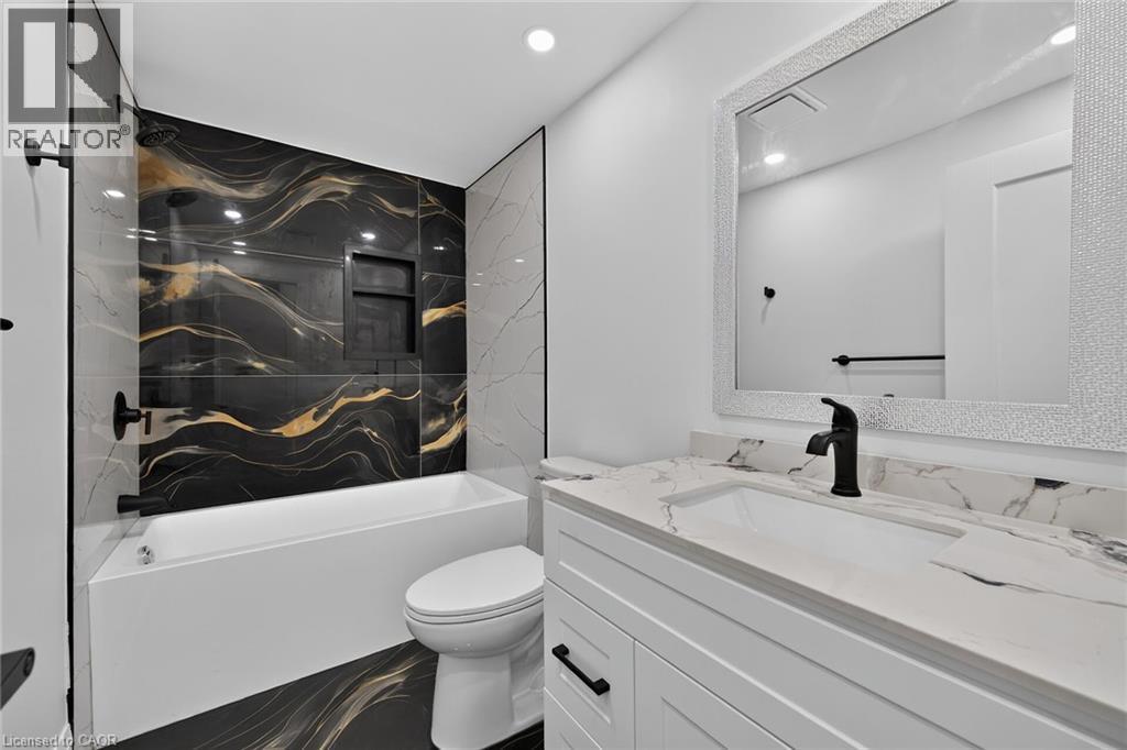 Built-in bathtub with a black and gold marbled tile surround, white vanity with a white countertop and integrated sink, matte black faucet, and a decorative framed mirror - 50 West 4Th Street, Hamilton, ON