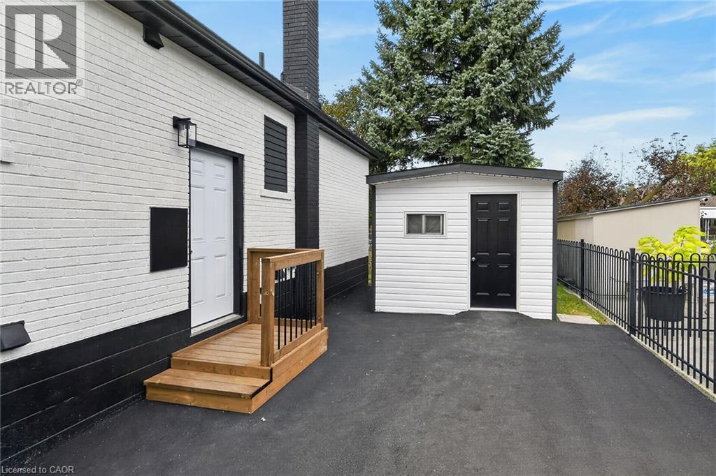 White brick exterior with black trim, wooden deck with railing, separate storage shed with white siding, black door, and small window, and a wrought iron fence - 50 West 4Th Street, Hamilton, ON