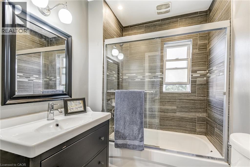 Vanity with integrated sink, framed mirror, wall-mounted light fixture, and tiled shower with glass enclosure - 11 Elm Street, Hamilton, ON