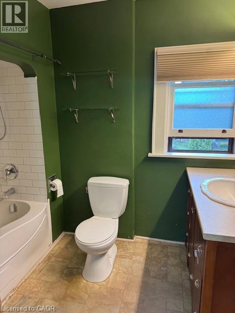 Bathroom featuring a white bathtub with subway tile surround, white toilet, wood-finish vanity with an integrated sink, and an exterior window with blinds - 255 Main Street, Cambridge, ON