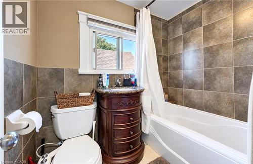 Tiled shower surround and wainscoting, white bathtub, wood-finish vanity with stone countertop, and window with trim - 3 Craigmiller Avenue, Hamilton, ON 