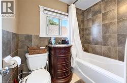 Tiled shower surround and wainscoting, white bathtub, wood-finish vanity with stone countertop, and window with trim -