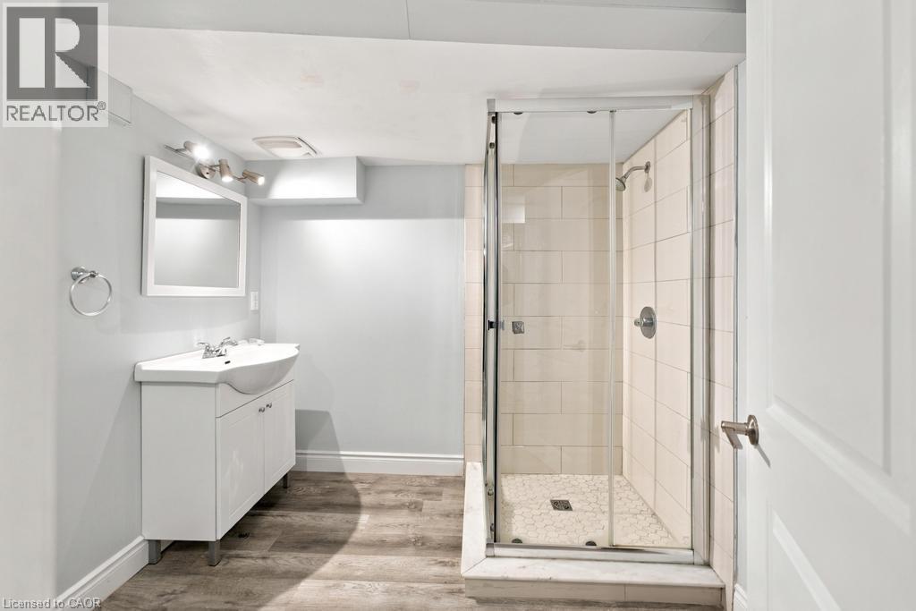 Walk-in shower with clear glass enclosure and subway tile surround, white vanity with integrated sink, wood-finish flooring, and wall-mounted vanity lighting - 284 East 21St Street, Hamilton, ON
