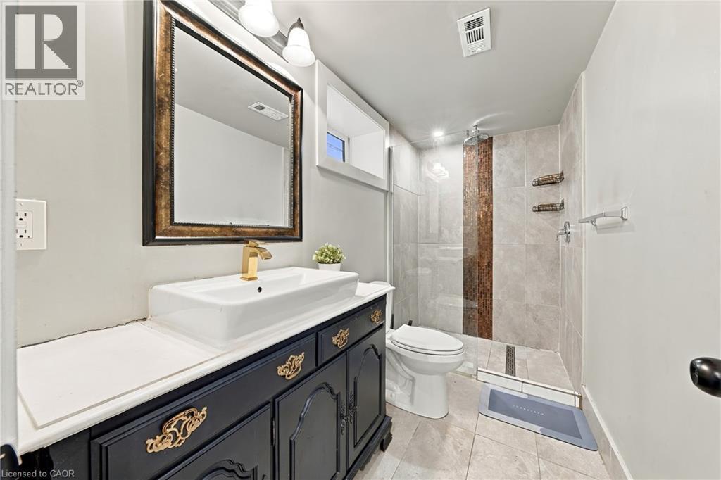 Vanity with a vessel sink and gold-finish faucet, framed mirror, tiled flooring, glass-enclosed shower with vertical accent tiling - 1231 Dunsmure Road, Hamilton, ON