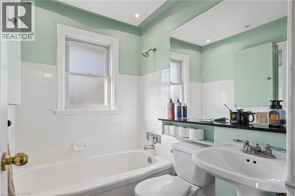 Full bathroom featuring a pedestal sink, white ceramic tiled walls, a bathtub with an integrated shower, and a mounted mirror - 366 East 17Th Street, Hamilton, ON