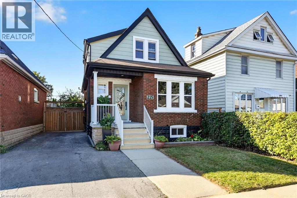 Brick and siding exterior with a peaked roofline, front porch, mature hedge, and paved driveway - 215 Paling Avenue, Hamilton, ON