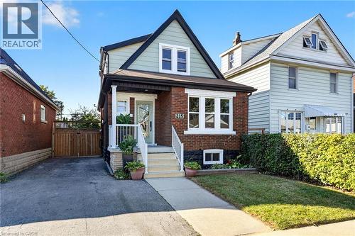 Brick and siding exterior with a peaked roofline, front porch, mature hedge, and paved driveway - 215 Paling Avenue, Hamilton, ON 