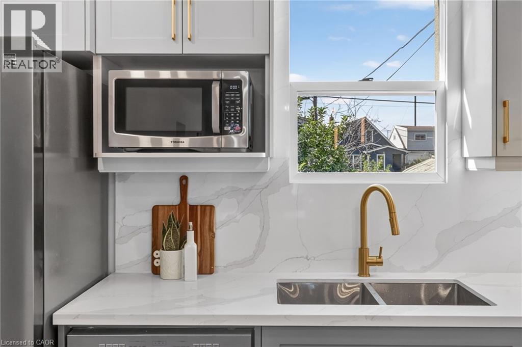 Stainless steel microwave, white shaker-style cabinetry with gold-tone hardware, white countertops, dual basin stainless steel sink, and a gold-tone gooseneck faucet - 215 Paling Avenue, Hamilton, ON